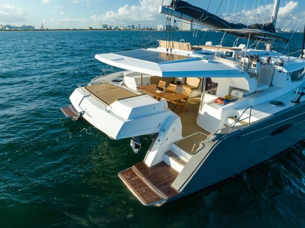 Luxury white catamaran yacht in blue coastal waters on a sunny day, featuring an open aft deck with teak swim platform, outdoor dining area and distant city skyline.