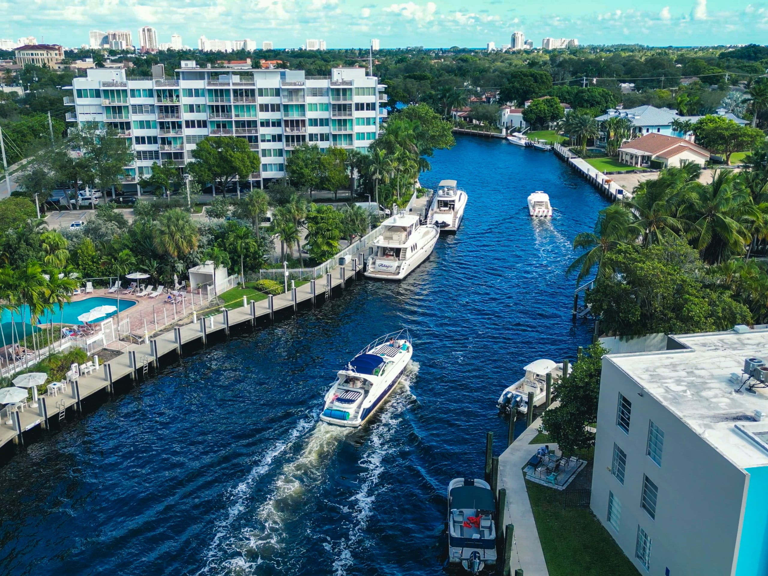 Aerial view of a sunny Florida waterfront canal with yachts cruising between palm-lined docks, waterfront condos and a pool