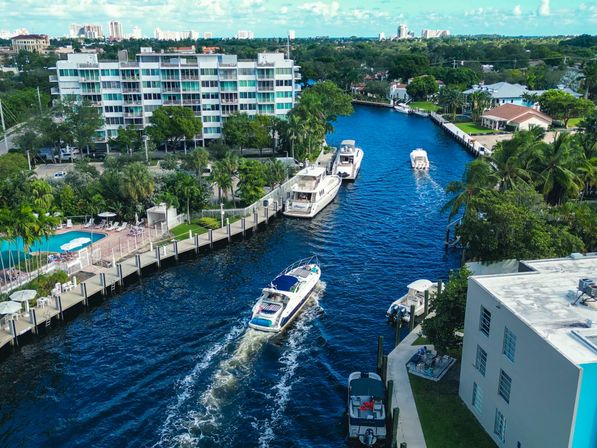 Aerial view of a sunny Florida waterfront canal with yachts cruising between palm-lined docks, waterfront condos and a pool