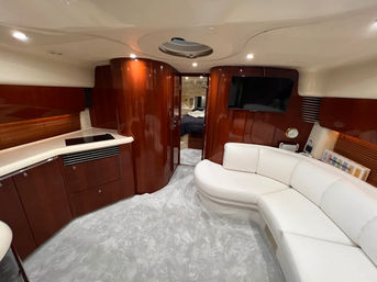 Luxurious yacht salon with curved white leather sofa, glossy cherry-wood cabinetry and galley cooktop, wall-mounted TV, plush light-gray carpet, and open doorway to a forward stateroom