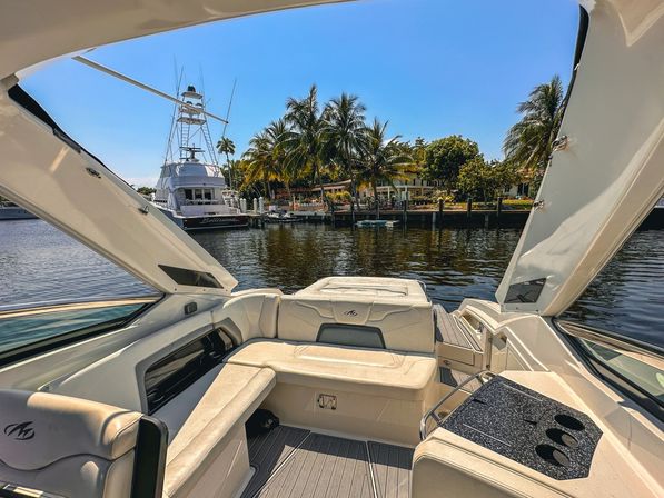View from a luxury motorboat’s open cockpit with cream cushioned seating and cup holders, looking out over a calm tropical marina with palm trees, waterfront homes and a large white sportfishing yacht under a bright blue sky.