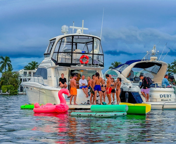 Boat party on a Miami waterfront: group of people socializing on a white yacht with a large pink flamingo float and colorful floating pads, palm trees and a cloudy sky