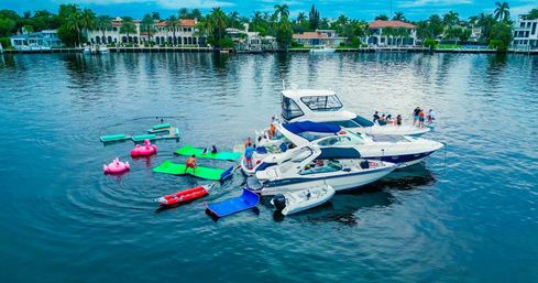 Aerial view of luxury yachts and boats in a tropical waterfront canal with bright green floating mats and pink flamingo inflatables, people socializing on decks, palm trees and upscale shoreline homes