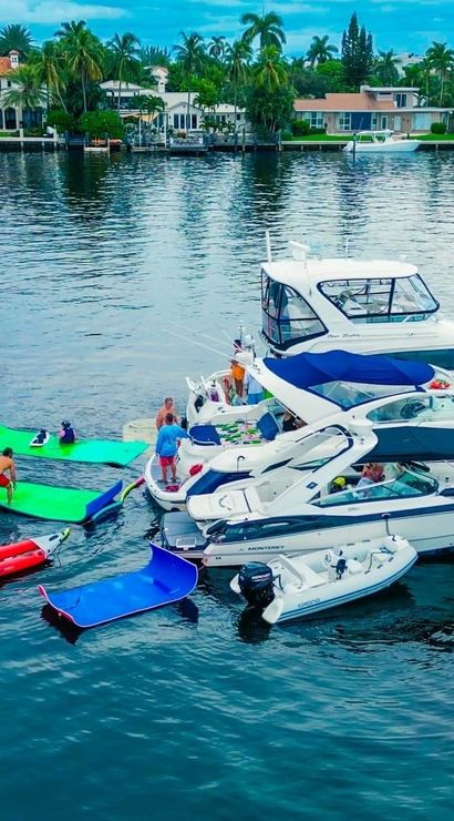 Aerial view of luxury yachts and boats in a tropical waterfront canal with bright green floating mats and pink flamingo inflatables, people socializing on decks, palm trees and upscale shoreline homes