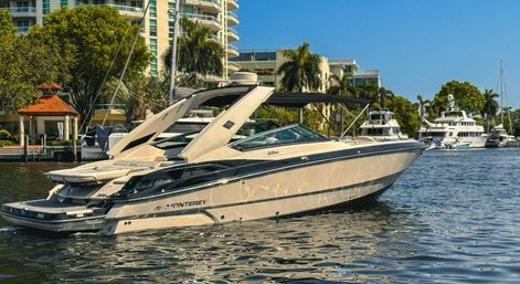 Sleek white luxury motorboat at a sunny marina, gliding on shimmering water past waterfront condos, palm trees, and moored yachts.