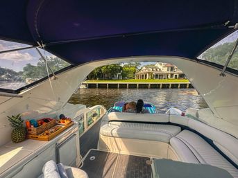 Sunlit view from a shaded luxury yacht cockpit with white leather seating and a snack tray (pineapple, fruit, pastries), two people relaxing at the stern on a striped float, facing a grand waterfront mansion across a calm river.