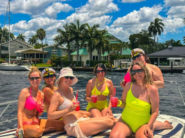Six women in colorful swimwear holding red cocktails and smiling on a boat in a sunny waterfront canal lined with palm trees, sailboats, and coastal homes.