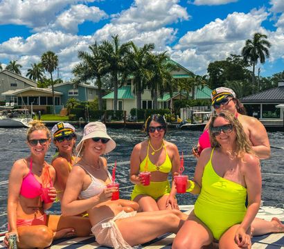 Six women in colorful swimwear holding red cocktails and smiling on a boat in a sunny waterfront canal lined with palm trees, sailboats, and coastal homes.