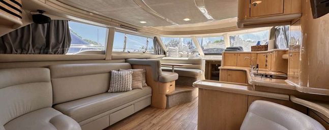 Sunlit luxury yacht interior salon with cream leather seating, wood floors, compact galley and cozy dinette by wraparound windows overlooking a marina.