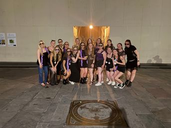 Group of young women in black and purple party outfits posing at night in an urban stone plaza, standing before a lit doorway and temporary wall, some wearing sunglasses and purple boas with a circular bronze plaza medallion in the foreground.