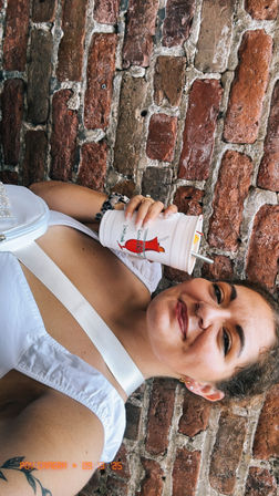 Smiling woman in a white tank top and crossbody bag leans against a rustic red brick wall holding a cold drink cup with a metal straw and red graphic — urban summer selfie showing a tattoo and septum ring.