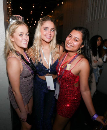 Three smiling women in sparkly party dresses and beaded necklaces pose for a friends' night out photo under string lights in a dim indoor nightclub, one wearing a 'Happy' tiara.