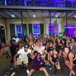 Large group photo of people cheering at a lively late-night street party on a historic porch with purple neon lights, outdoor downtown nightlife scene