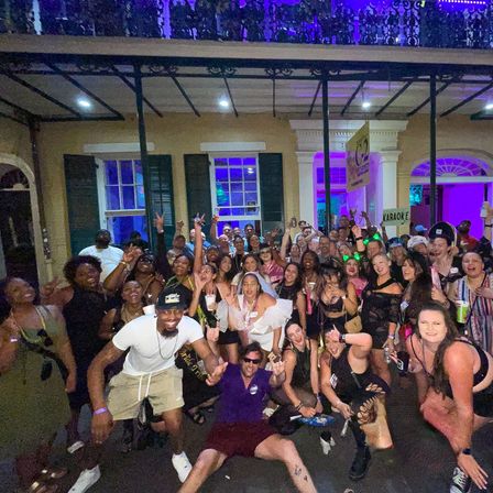 Large group photo of people cheering at a lively late-night street party on a historic porch with purple neon lights, outdoor downtown nightlife scene