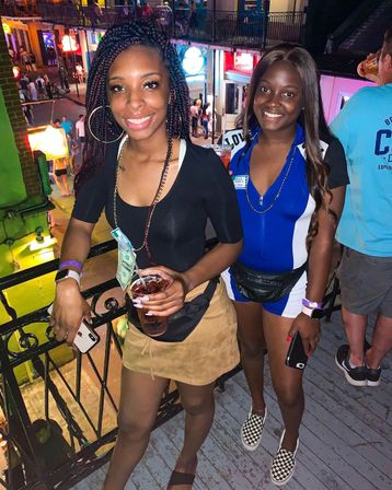 Two smiling women on a balcony overlooking a busy neon-lit downtown entertainment street at night, one holding a drink with dollar bills on a bead necklace, both wearing casual night-out outfits and fanny packs.