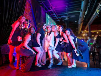 Group of eight women posing and smiling on a neon-lit bar stage during a lively night out, colorful purple and pink club lighting and party outfits in an indoor nightlife scene.