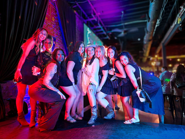 Group of eight women posing and smiling on a neon-lit bar stage during a lively night out, colorful purple and pink club lighting and party outfits in an indoor nightlife scene.