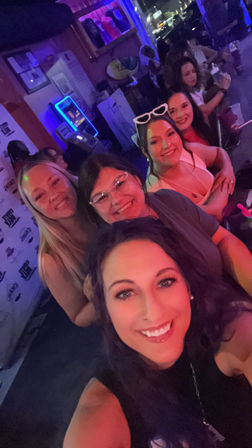 Group selfie of five smiling women enjoying a neon-lit bar or nightclub with purple-pink lighting and drinks on the counter