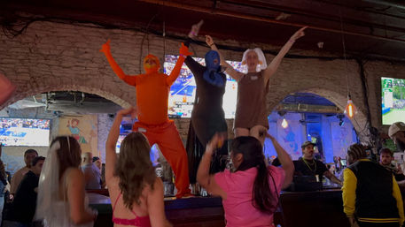 Three costumed performers in orange, blue and brown outfits dancing on a bar counter as a cheering crowd watches inside a dim sports bar with exposed brick walls and TVs showing games.