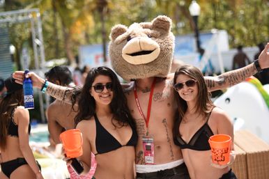 Sunny outdoor pool party scene: two women in black bikinis holding orange buckets and a tattooed man wearing a large fuzzy bear-head mascot, posing poolside with drinks and smiles.