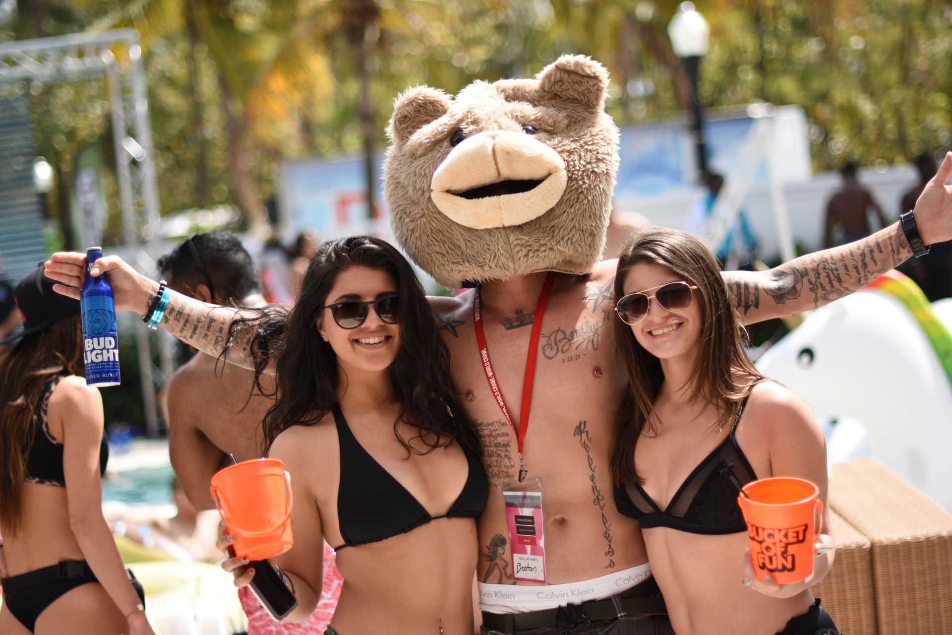 Sunny outdoor pool party scene: two women in black bikinis holding orange buckets and a tattooed man wearing a large fuzzy bear-head mascot, posing poolside with drinks and smiles.