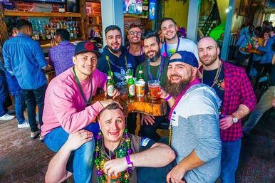 Group of friends at a lively bar wearing colorful Mardi Gras beads, laughing and holding beers around a wooden high-top table with a crowded bar interior in the background.