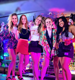 Five friends posing and smiling on a neon-lit bar counter at a lively nightlife spot, wearing colorful summer outfits and holding drinks beneath string lights and hanging bras.