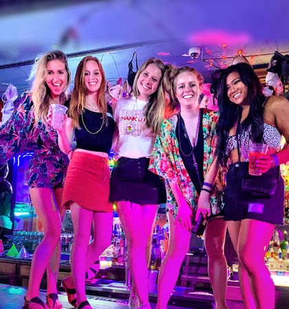 Five friends posing and smiling on a neon-lit bar counter at a lively nightlife spot, wearing colorful summer outfits and holding drinks beneath string lights and hanging bras.