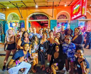 Festive group of friends posing for a neon-lit nightlife street photo outside colorful shopfronts, many wearing beads and holding drinks.