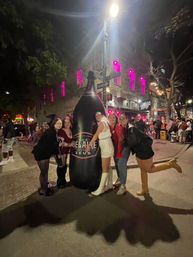 Group of five people hugging an oversized inflatable champagne bottle on a busy downtown nightlife street at night, pink-lit building windows and crowds in the background.