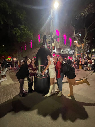 Group of five people hugging an oversized inflatable champagne bottle on a busy downtown nightlife street at night, pink-lit building windows and crowds in the background.