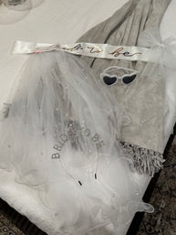 White pearl-studded bridal veil, 'bride-to-be' satin sash, rhinestone headband and heart-shaped sunglasses on a gray blanket — bachelorette party accessories