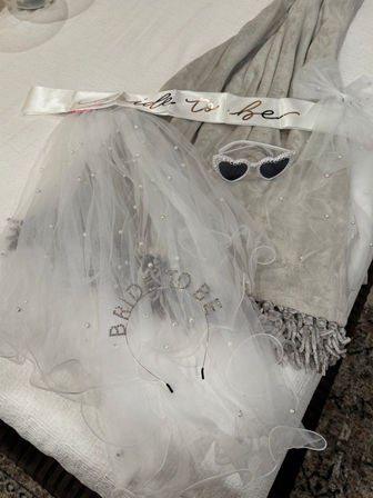 White pearl-studded bridal veil, 'bride-to-be' satin sash, rhinestone headband and heart-shaped sunglasses on a gray blanket — bachelorette party accessories
