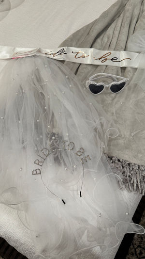 White pearl-studded bridal veil, 'bride-to-be' satin sash, rhinestone headband and heart-shaped sunglasses on a gray blanket — bachelorette party accessories