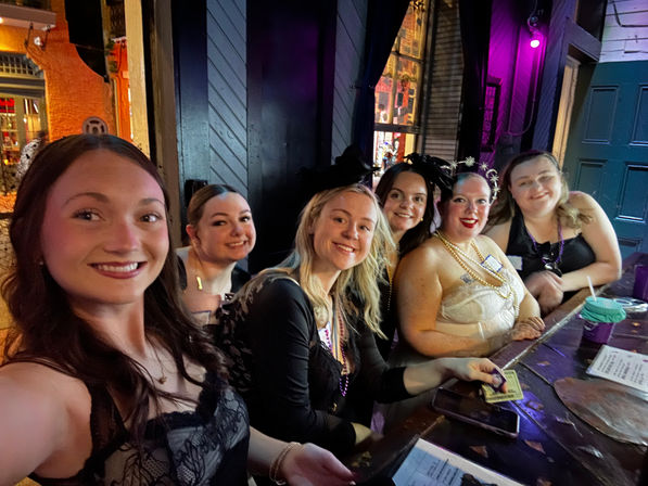 Selfie of six smiling women seated at a neon-lit bar counter wearing party outfits and beaded necklaces, enjoying a festive night out