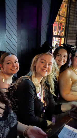 Selfie of six smiling women seated at a neon-lit bar counter wearing party outfits and beaded necklaces, enjoying a festive night out