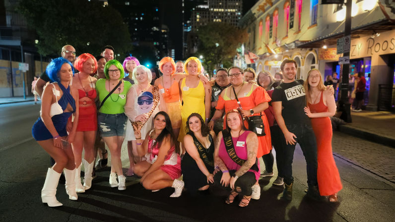 Group of friends in colorful wigs and costumes posing for a celebratory photo on a lively downtown bar-lined street at night