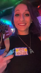 Nightlife selfie of a smiling person in a black top at a neon-lit bar, holding a beer and a blue prop, wearing a playful name sticker on their chest.