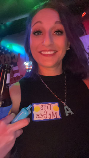 Nightlife selfie of a smiling person in a black top at a neon-lit bar, holding a beer and a blue prop, wearing a playful name sticker on their chest.