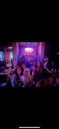 Bachelorette party group of eight friends smiling and posing in a purple-lit bar under a decorative neon sign, wearing party sashes and leopard-print outfits with drinks on a nearby table.