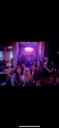 Bachelorette party group of eight friends smiling and posing in a purple-lit bar under a decorative neon sign, wearing party sashes and leopard-print outfits with drinks on a nearby table.