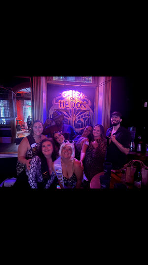 Bachelorette party group of eight friends smiling and posing in a purple-lit bar under a decorative neon sign, wearing party sashes and leopard-print outfits with drinks on a nearby table.