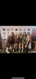 Six women wearing leopard-print outfits and sashes posing together in front of a branded step-and-repeat backdrop inside a New Orleans bar for a bachelorette-style celebration.