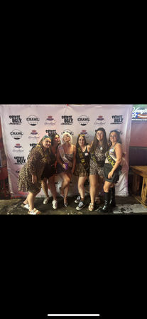 Six women wearing leopard-print outfits and sashes posing together in front of a branded step-and-repeat backdrop inside a New Orleans bar for a bachelorette-style celebration.