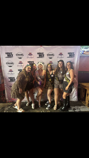 Six women wearing leopard-print outfits and sashes posing together in front of a branded step-and-repeat backdrop inside a New Orleans bar for a bachelorette-style celebration.