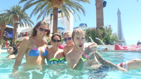 Four friends smiling in a sunny Las Vegas pool party, palm trees overhead and an Eiffel Tower–style hotel tower in the background