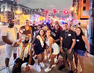 Lively group of friends posing on a busy downtown nightlife street at night, smiling and celebrating with neon lights, traffic signals, and one person wearing a sash.