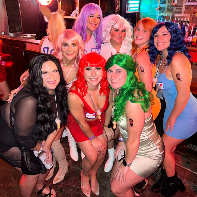 Group of eight friends in colorful wigs and party dresses smiling for a lively bar group photo under neon lights, playful temporary tattoos and festive nightlife vibes