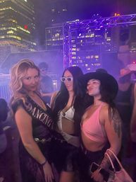 Three friends posing at a rooftop party at night under purple stage lights with lit city skyscrapers in the background, one wearing a sash and another in a pink top, festive and smiling.