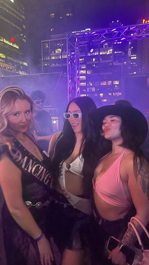 Three friends posing at a rooftop party at night under purple stage lights with lit city skyscrapers in the background, one wearing a sash and another in a pink top, festive and smiling.